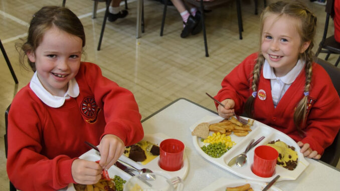 Two girls are sitting at the table in school having hot lunch and smiling.