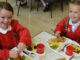 Two girls are sitting at the table in school having hot lunch and smiling.