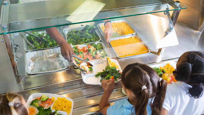 Two school children are seen at the school lunch counter. Someone is serving salad to one of the girls. The other girl has taken her share and is standing nearby. A third girl is also seen in the background with her plate filled.