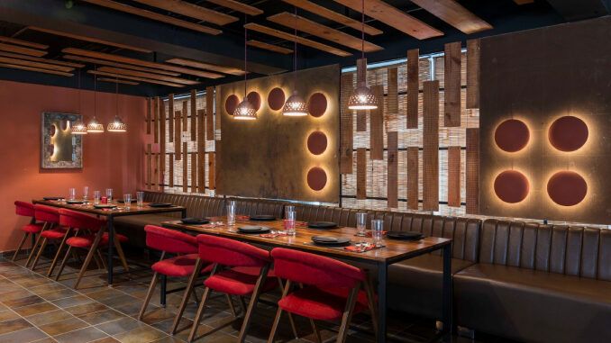 Tables and chairs are arranged with plates, spoons and glasses in a restaurant that is brightened with lantern light setting.